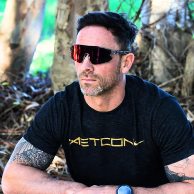 Man wearing sunglasses and a black t-shirt with 'Aetcon' logo in a forest setting